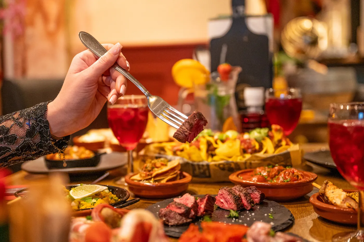 Een gast prikt een stukje biefstuk van een tafel vol tapas, met sangria en cocktails