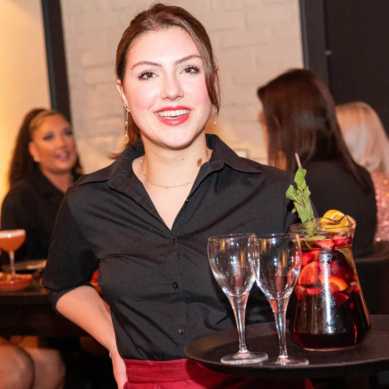 Een lachende gastvrouw brengt een kan rode sangria met glazen naar tafel
